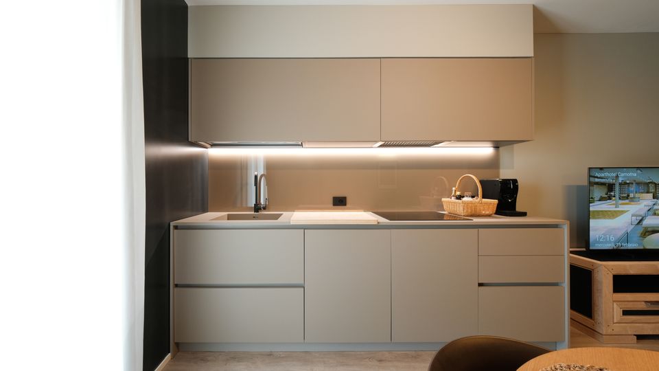Cucina moderna lineare con mobili grigio opaco, pensili sospesi e illuminazione a LED sotto pensile. Piano di lavoro chiaro con lavello e piano cottura a induzione, pareti neutre e pavimento effetto legno; a destra si intravede un mobile TV in legno.
