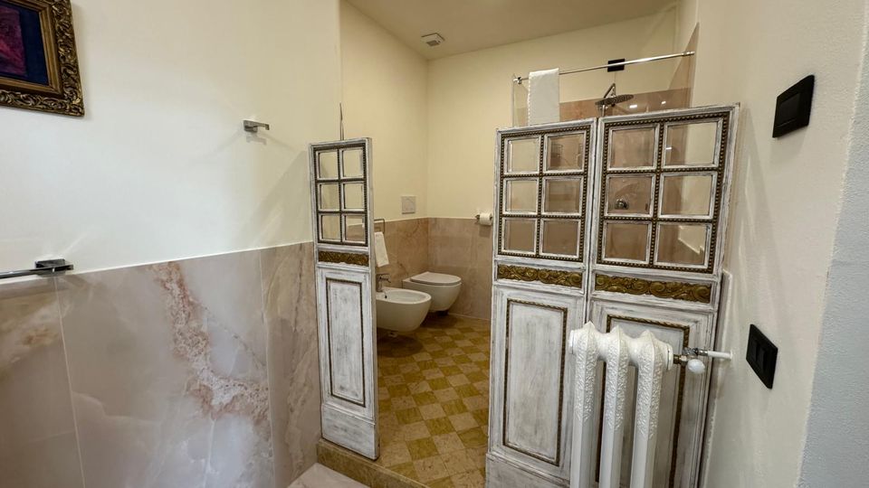Bagno interno con pareti bianche e rivestimento in piastrelle effetto marmo, pavimento a quadri beige e sanitari sospesi sul fondo. L’area doccia è separata da pannelli decorativi in legno bianco con riquadri a vetro e dettagli dorati, con radiatore bianco in primo piano.