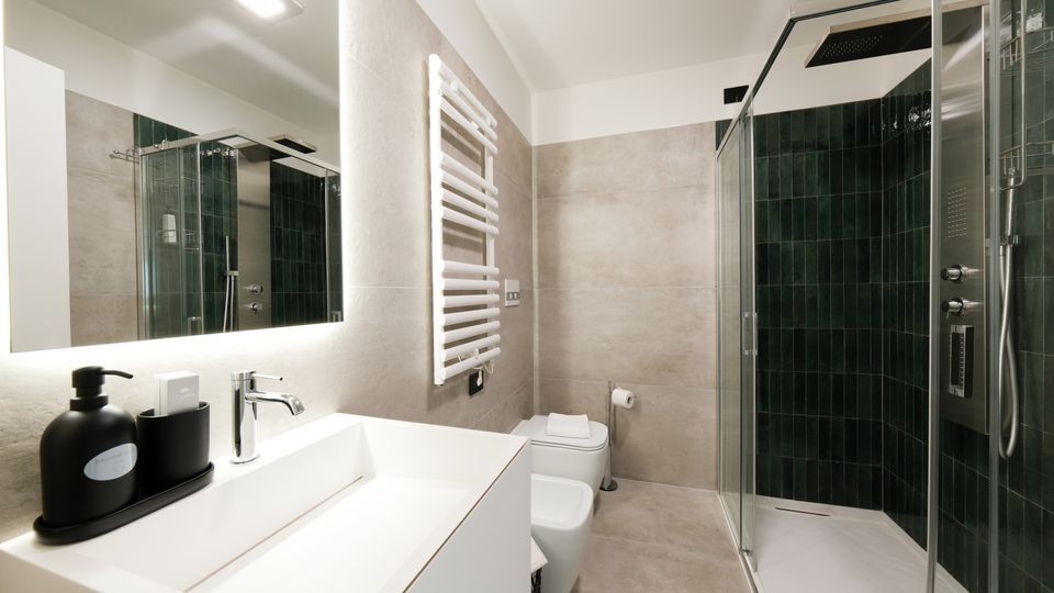 Bagno moderno con lavabo bianco e grande specchio illuminato, pareti rivestite in piastrelle grigie e scaldasalviette verticale. A destra una doccia con box in vetro e rivestimento in piastrelle verde scuro, con soffione a pioggia.