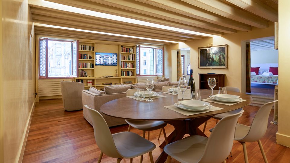 Ampio soggiorno-pranzo in un appartamento con pavimento in legno e soffitto con travi a vista, con tavolo rotondo apparecchiato e sedie moderne in primo piano. Sullo sfondo ci sono divani, librerie integrate e grandi finestre, con un passaggio che conduce a una camera da letto.