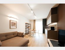 Open space moderno con soggiorno e cucina lineare a vista: divano beige, pavimento in legno chiaro e tavolo rotondo con sedie vicino a una grande finestra con tende. Pensili scuri e bianchi illuminati da luci a binario e una scaffalatura metallica completano l’ambiente.