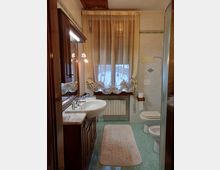 Bagno stretto con pavimento verde e pareti piastrellate chiare, mobile lavabo in legno con specchiera illuminata. In fondo una finestra con tende leggere e termosifone sotto, e sul lato destro WC e bidet accanto a una doccia con parete in vetro.