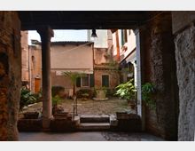 Piccolo cortile interno in un contesto urbano storico, pavimentato in mattoni e pietra, con un pozzo o vasca al centro e numerose piante in vaso. L’inquadratura è sotto un portico con colonne e travi a vista, circondato da facciate intonacate e muri in mattoni con finestre e persiane.