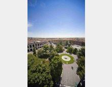 Veduta aerea di una grande piazza urbana con alberi e viali pedonali, con una fontana circolare al centro. Sullo sfondo si vedono edifici storici, tra cui un’ampia struttura ad arcate simile a un anfiteatro e un palazzo con facciata classica.