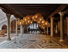 Cortile porticato in un edificio storico con colonne in pietra, soffitto a travi lignee e pavimento a mosaico geometrico. Al centro è sospesa una grande installazione luminosa ramificata, con archi in mattoni e finestre sullo sfondo.