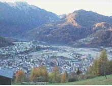 Vista panoramica di una valle alpina con un paese esteso tra campi e zone agricole, circondato da montagne boscose. In primo piano si vede un pendio erboso con alberi autunnali e alcune costruzioni ai margini.