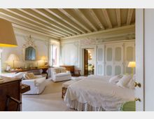 Camera da letto in stile classico con soffitto a travi dipinte e pareti decorate a motivi floreali pastello. Letto matrimoniale con biancheria chiara, armadi a pannelli, specchio ornamentale e area salotto con poltrone vicino a una grande finestra.