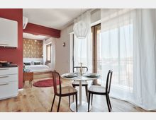 Interno di un monolocale moderno con zona pranzo con tavolo rotondo e sedie, cucina a vista e grande portafinestra con tende bianche che dà su un balcone. Sul fondo si vede la zona notte rialzata con letto, parete decorata e pavimento in legno chiaro.