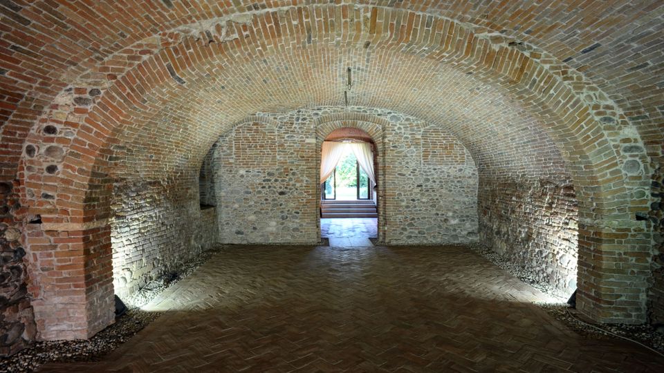 Interno di una cantina con volte a botte in mattoni e pareti in pietra grezza, con un pavimento in cotto disposto a spina di pesce. Sullo sfondo, una porta ad arco con tende leggere lascia intravedere un giardino illuminato.