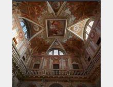 Interno di un palazzo storico con soffitto a volta decorato da affreschi e riquadro centrale dipinto, circondato da lunette. Pareti con balconate in pietra e grandi finestre ad arco che illuminano l’ambiente.