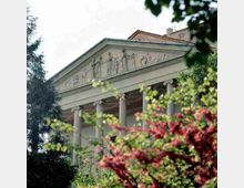 Facciata di un edificio in stile neoclassico con frontone triangolare decorato a bassorilievo e un portico sorretto da alte colonne. L’edificio è parzialmente nascosto da alberi e vegetazione in primo piano.