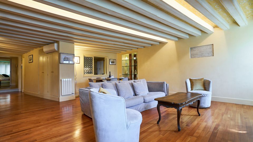 Soggiorno open space con pavimento in parquet e soffitto a travi in legno a vista dipinte di chiaro, con pareti color crema. Al centro ci sono un divano e poltrone chiare attorno a un tavolino, con zona pranzo e corridoio sullo sfondo.