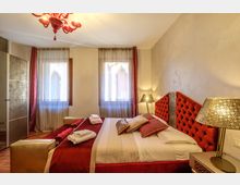 Camera da letto in stile hotel con letto matrimoniale, testiera imbottita rossa e comodini con lampade traforate. Due finestre con tende leggere e drappeggi rossi illuminano la stanza, con pavimento in legno e lampadario in vetro rosso al soffitto.