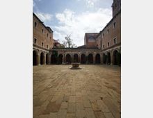 Cortile interno lastricato in pietra, circondato su tre lati da portici ad archi su colonne e da edifici in mattoni con finestre rettangolari. Al centro si trova una piccola fontana circolare con una pianta, sotto un cielo parzialmente nuvoloso.