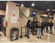 Atrio interno di un cinema o teatro con scale in pietra che salgono al piano superiore, soffitto a griglia con luci incassate e cartellone digitale con numeri delle sale. Pareti beige con segnaletica e piantina, estintore e poster, oltre a ingresso vetrato e colonne nell’area laterale.