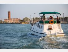 Imbarcazione a motore bianca con tettuccio verde che naviga in un porto turistico, lasciando una scia sull’acqua. Sullo sfondo si vedono numerosi alberi di barche a vela, un lungo edificio con torre in mattoni e pali di illuminazione lungo la riva.