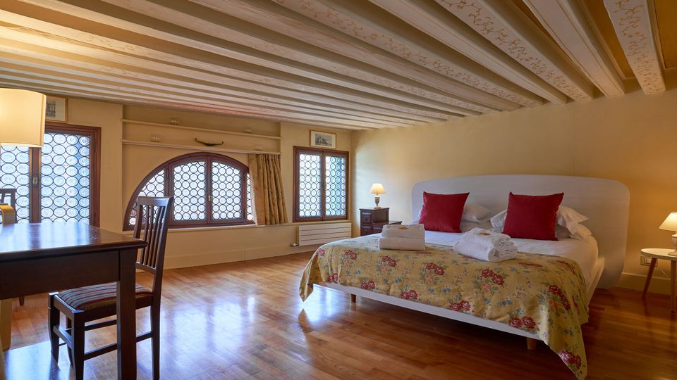 Camera da letto in stile tradizionale con pavimento in legno, soffitto a travi dipinte e ampie finestre ad arco con vetri decorati. Al centro c’è un letto matrimoniale con copriletto floreale e cuscini rossi, affiancato da comodini con lampade, e un tavolo con sedie vicino alla finestra.