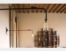 Interno con soffitto a travi in legno e pareti chiare, illuminato da lampade a braccio in metallo nero con lampadine a vista. Sullo sfondo si vede una libreria a parete in legno dalla forma organica con ripiani irregolari.