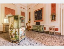 Sala d’interni in stile storico con pareti bianche e riquadri rosso mattone, pavimento in graniglia e arredi antichi. In primo piano c’è una grande alcova paravento dipinta, accanto a un’arpa, una credenza decorata, specchi con cornici dorate e una sedia imbottita.