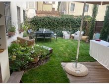 Giardino privato con prato curato, aiuole fiorite e zona pranzo con tavolo e sedie vicino a siepi e recinzioni. In primo piano una pedana in legno con ombrellone e un tavolo coperto da tovaglia bianca.