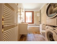 Bagno moderno e stretto con piastrelle beige, pavimento effetto legno e vasca con doccia sotto una finestra con vetro opaco. A sinistra ci sono un grande termoarredo e un mobile lavabo sospeso, mentre a destra sono presenti WC e colonna con lavatrice e asciugatrice.