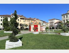 Piazza con prato curato e aiuole in grandi fioriere bianche, con una piccola fontana e percorsi pavimentati. Al centro c’è un padiglione decorativo rosso e bianco in stile giostra, circondato da edifici storici con portici e tetti in coppi.