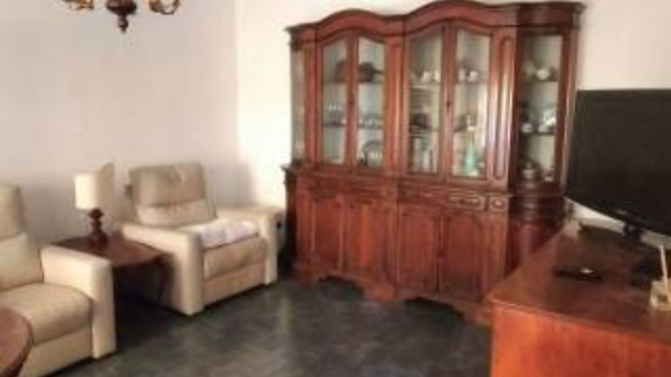 Soggiorno con due poltrone beige e un tavolino con lampada, pavimento scuro e pareti chiare. Sul fondo c’è una grande vetrina in legno con ante in vetro e, a destra, un mobile in legno con televisore; dal soffitto pende un lampadario classico.