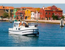 Canale lagunare con un’imbarcazione bianca in primo piano e, sullo sfondo, una fila di case colorate con tetti in tegole rosse lungo la riva. Si vedono pali d’ormeggio in legno e un lungomare con lampioni davanti alle abitazioni.