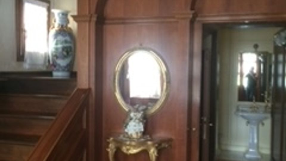 Interno di un'abitazione con pareti rivestite in legno scuro, un arco decorativo, uno specchio con cornice dorata e consolle sottostante. A sinistra si trova una scala in legno, e sullo sfondo è visibile un bagno con lavabo bianco.