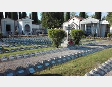 Cimitero con file ordinate di piccole lapidi basse in pietra su prato e ghiaia, disposte in settori rettangolari. Sullo sfondo si vedono cappelle funerarie bianche con croci e cipressi alti che delimitano l’area.