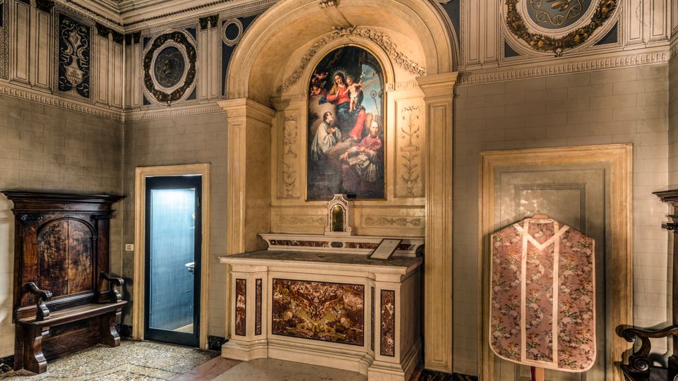 Interno di una piccola cappella con altare in marmo sotto un arco e grande dipinto religioso sopra, circondata da pareti decorate con stucchi e medaglioni sul soffitto. A lato si vedono panche e arredi in legno, una porta vetrata e un paramento liturgico esposto in una nicchia.