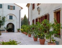 Cortile interno pavimentato tra edifici in intonaco chiaro, con finestre e portoni in legno scuro e un arco d’ingresso sul lato sinistro. Lungo il muro di destra si allineano grandi vasi in terracotta con alberi di agrumi, con cipressi sullo sfondo.