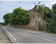 Strada asfaltata di campagna che costeggia un alto muro in mattoni e un edificio in rovina con piccole finestre ad arco. La struttura è parzialmente coperta da edera e alberi fitti, con un cancello metallico e un lampione vicino all’angolo.