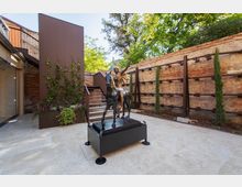 Cortile interno pavimentato con statue in bronzo su piedistallo al centro, circondato da muri in mattoni a vista e strutture metalliche moderne. Sullo sfondo si vedono scale, fioriere con rampicanti e alberi che ombreggiano lo spazio.