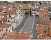 Veduta aerea di una piazza rettangolare in un centro storico, circondata da edifici con tetti in coppi rossi. Sul lato sinistro si distingue una grande chiesa in pietra con facciata classica e campanile, mentre la piazza è attraversata da un percorso centrale con lampioni e un monumento.