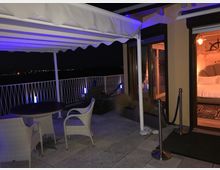 Terrazza esterna notturna con tavolo e sedie in vimini sotto una tenda retrattile, affacciata su un panorama buio con luci in lontananza oltre la ringhiera. A destra si vedono porte vetrate illuminate che danno su un interno con camera da letto.
