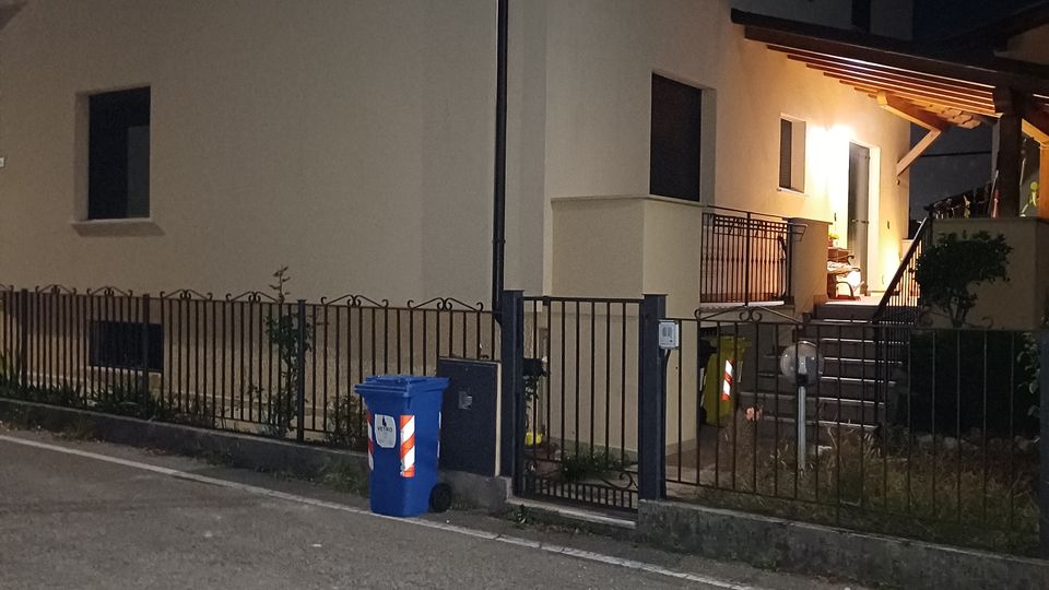 Esterno notturno di una casa a due piani con facciata chiara e finestre scure, circondata da una recinzione metallica con cancello. A destra si vede l’ingresso illuminato con piccolo portico in legno e una scalinata che sale dal marciapiede.