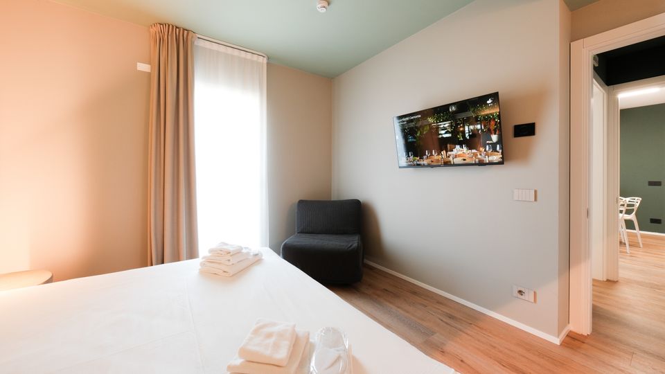 Camera da letto moderna con letto matrimoniale, pavimento in legno e pareti dai toni chiari; una grande finestra con tende lascia entrare luce naturale. Sulla parete è montata una TV, accanto a una poltrona scura e a una porta che conduce a un’altra stanza.