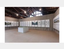 Interno di una sala espositiva con pavimento in cemento e soffitto alto in legno con travi a vista e lucernari. Alle pareti sono appesi pannelli con disegni e fotografie, mentre al centro si trova un basamento bianco con un plastico.