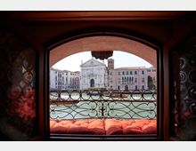 Vista dal’interno di una finestra ad arco con ringhiera in ferro battuto e cuscini rossi, affacciata su un canale con imbarcazioni. Di fronte si vedono la facciata di una chiesa barocca e palazzi storici lungo l’acqua.