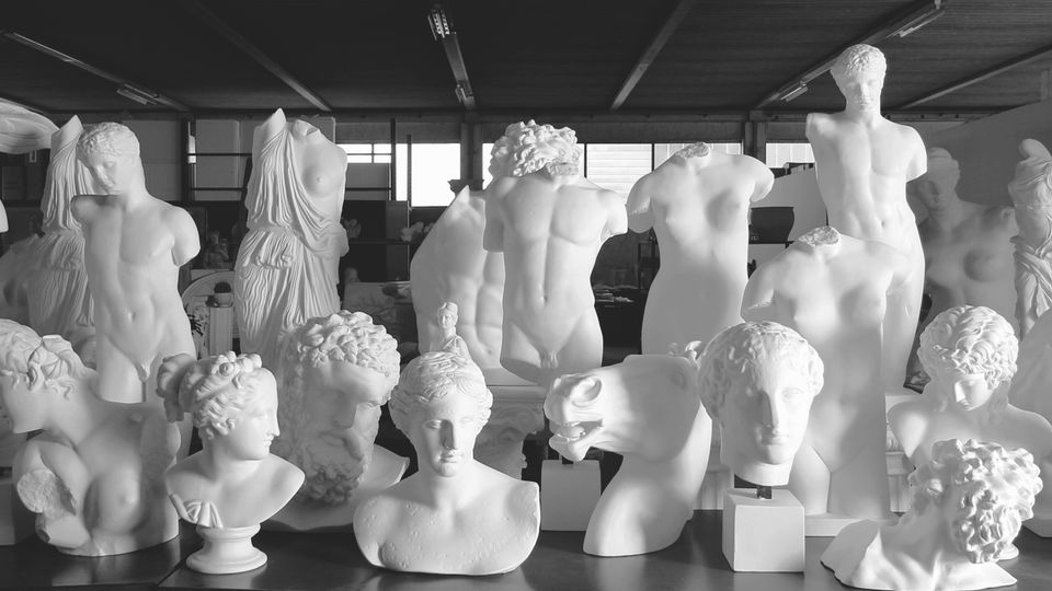 Interno di un laboratorio o magazzino d’arte con numerose statue e busti bianchi in gesso o marmo disposti su tavoli e piedistalli. L’ambiente ha soffitto industriale e finestre sullo sfondo che illuminano le sculture classiche.