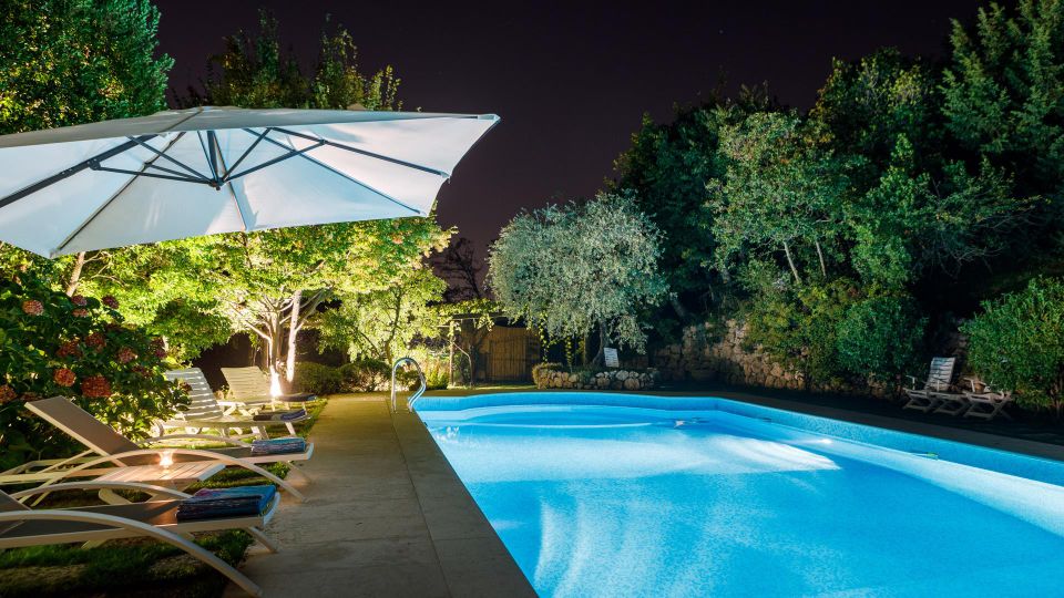 Piscina all’aperto illuminata di notte, con acqua azzurra e bordo in cemento. Lungo il lato ci sono lettini prendisole sotto un grande ombrellone bianco, circondati da alberi e vegetazione fitta.