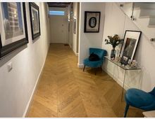 Corridoio interno di un’abitazione con pavimento in parquet a spina di pesce e pareti bianche, decorato con quadri incorniciati. A destra ci sono una consolle con specchio, oggetti e fiori, due sedie imbottite turchesi e una scala con gradini bianchi.