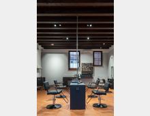 Interno di un salone da parrucchiere con pavimento in cotto e soffitto a travi di legno a vista con faretti. Al centro ci sono postazioni con specchi e sedute nere, mentre sul fondo si vedono finestre con inferriate e una parete con espositori di prodotti.