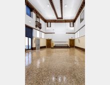 Ampia sala interna vuota con pavimento in terrazzo lucido, pareti bianche e cornici in legno scuro, soffitto alto con travi decorative e illuminazione a plafoniere. Sul fondo c’è un piccolo palco con tavolo, affiancato da porte in legno e finestre alte con tende scure.