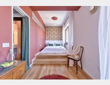 Camera da letto luminosa con letto su pedana rialzata e parete di fondo con carta da parati decorata e testiera bianca. Pavimento in legno, pareti rosa, finestra con tende bianche e accesso al bagno tramite apertura con tenda sul lato sinistro.