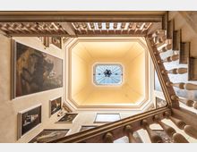 Veduta dal basso di un vano scala interno con ringhiere in pietra e pareti decorate da grandi dipinti incorniciati. Al centro si apre un lucernario ottagonale con vetrata e lampadario, incorniciato da un soffitto a riquadri color crema.