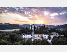 Veduta aerea di una villa storica bianca su una collina boscosa, con lunga scalinata centrale e cipressi che conducono all’edificio con cupola. Sullo sfondo si vedono un fiume o lago, una valle e colline montuose sotto un cielo al tramonto.