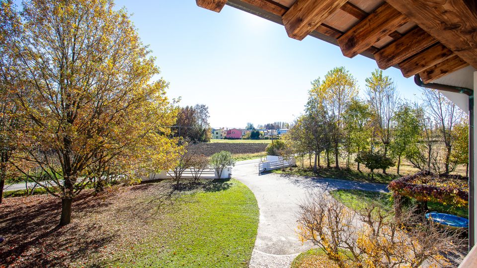 Vista da un portico con travi in legno su un giardino con prato, vialetto curvo e cancello bianco, circondato da alberi. Sullo sfondo si vedono campi coltivati e alcune case in una zona rurale.