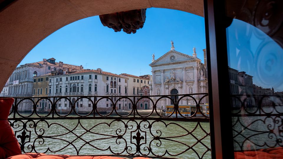 Vista da una terrazza coperta con ringhiera in ferro battuto sul Canal Grande, con facciate di palazzi storici e una chiesa monumentale in pietra sull’altra sponda. In primo piano si vedono cuscini rossi e una vetrata laterale che riflette parte della scena.
